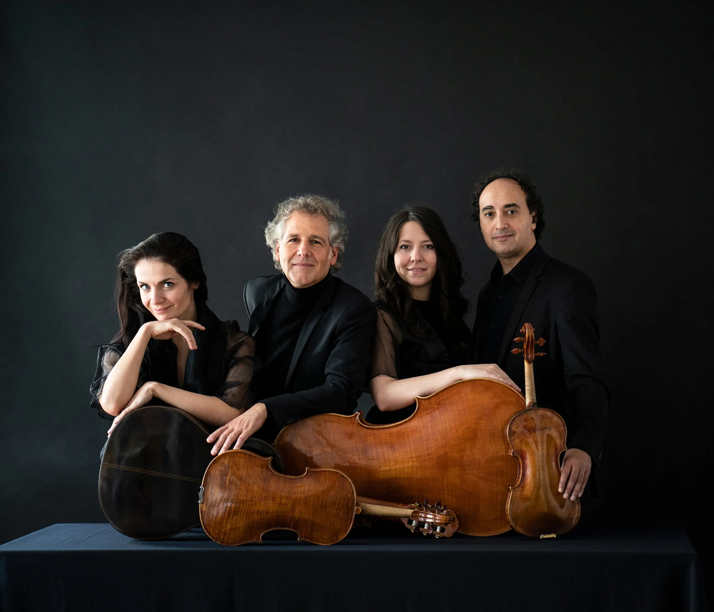 The four musicians standing behind a table on which a cello, a guitar, a viola and a violin are laid out