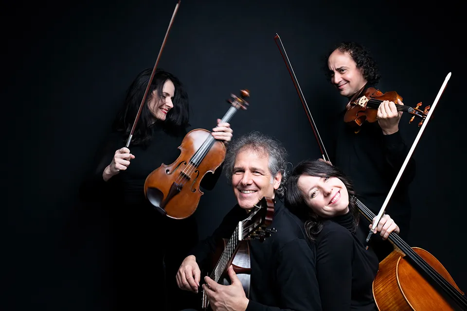 The four musicians in black against a dark studio background, the violist and violinist standing with raised bows behind the seated guitarist and cellist