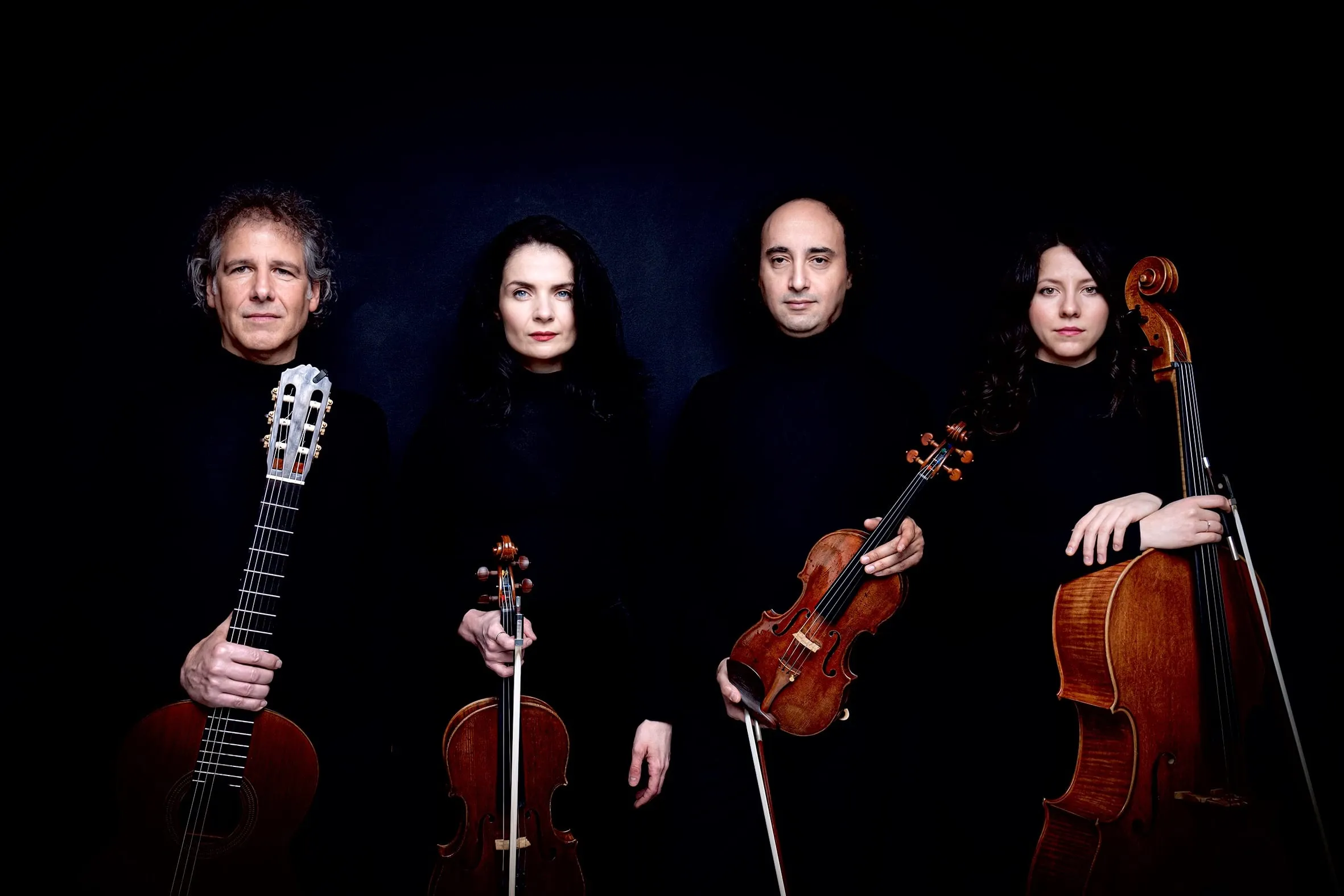 Group portrait of the four musicians with their instruments, lit warmly against black