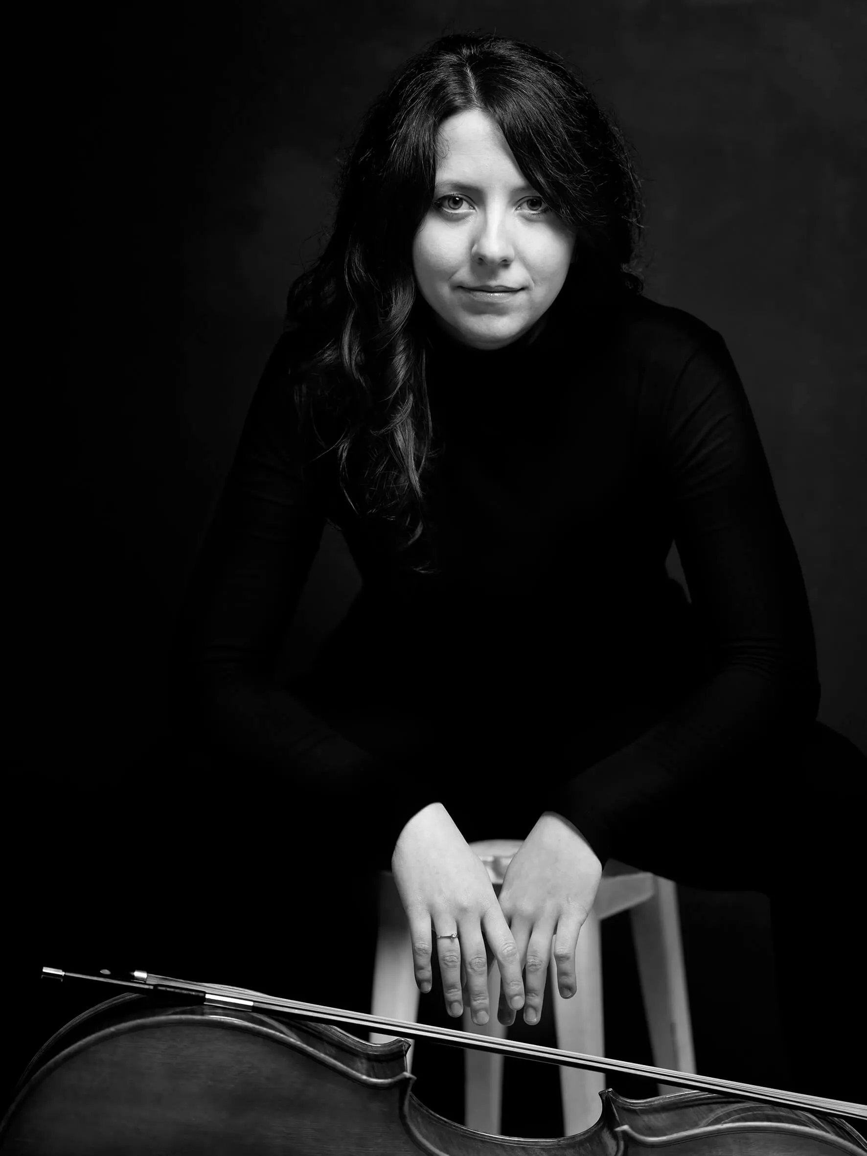 Liliana Kehayova looking into the camera, her hands resting on the top of her cello, black-and-white portrait