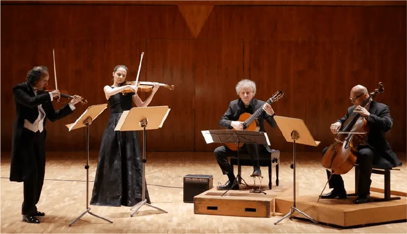 The ensemble performing on a wooden concert stage: violinist and violist standing, guitarist and cellist seated